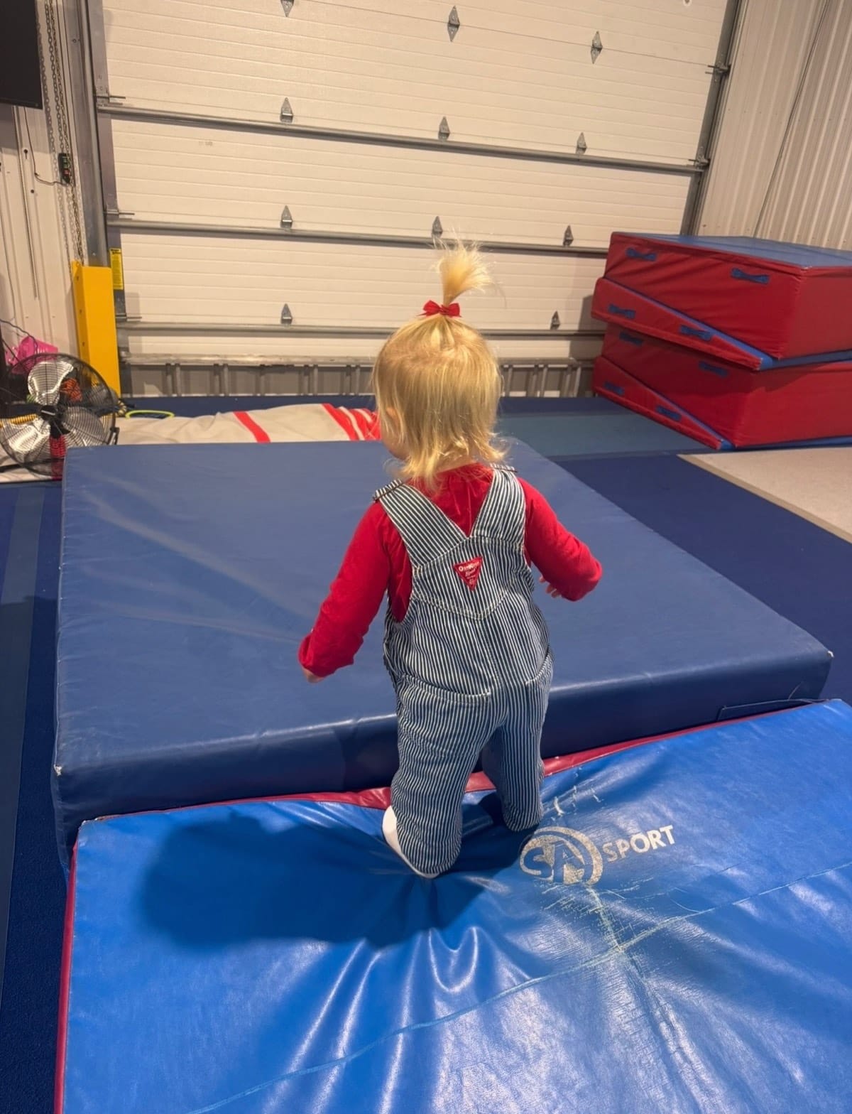 A toddler in red and striped overalls climbing onto a stack of blue mats at CheerCore Jags