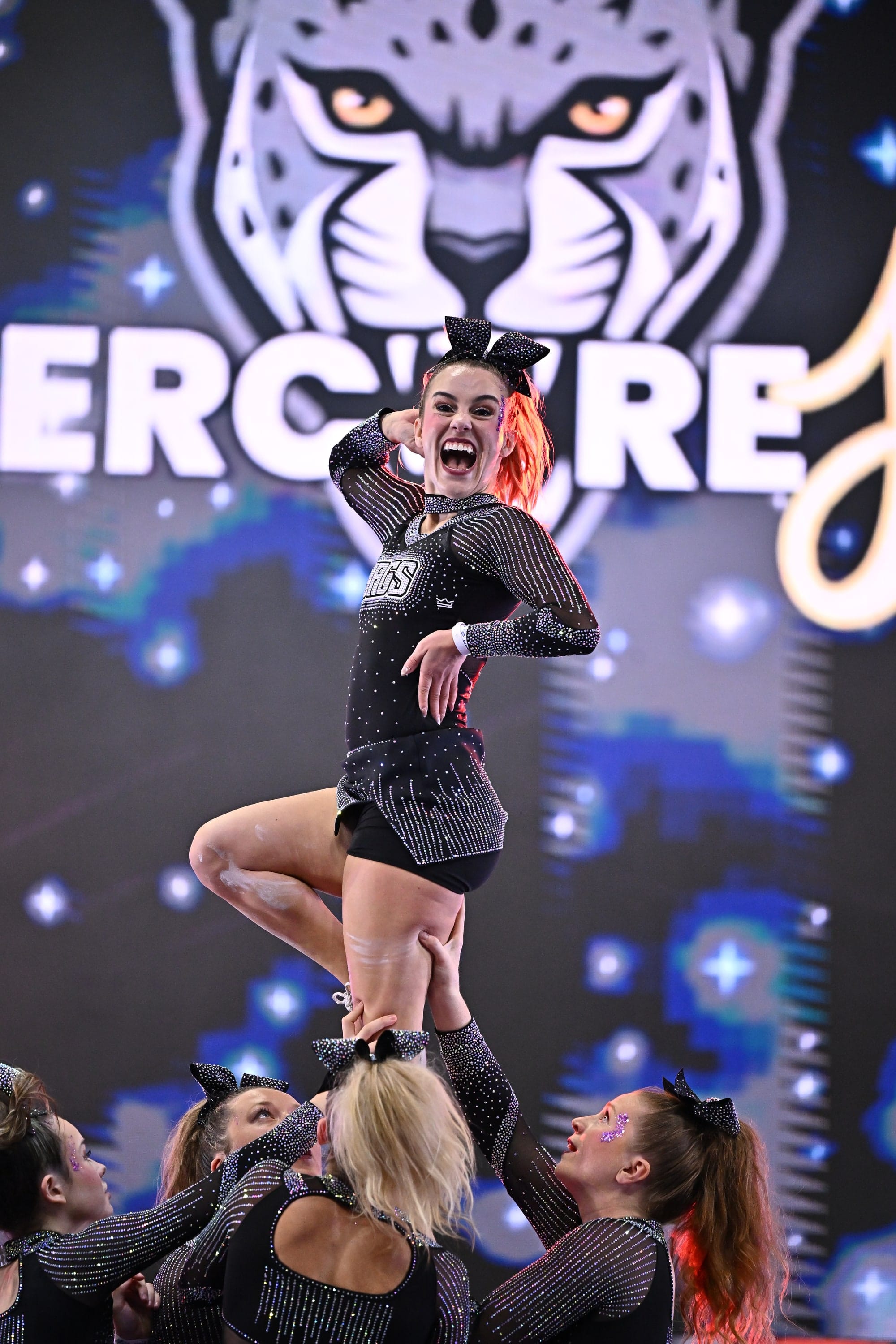 An Elite Cheer flyer held overhead in a stunt in front of the CheerCore Jags backdrop